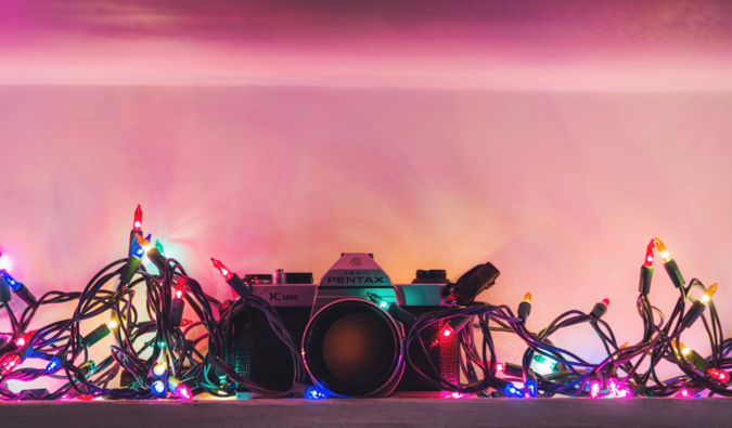 A small digital camera wrapped in Christmas lights in front of a bright pink background