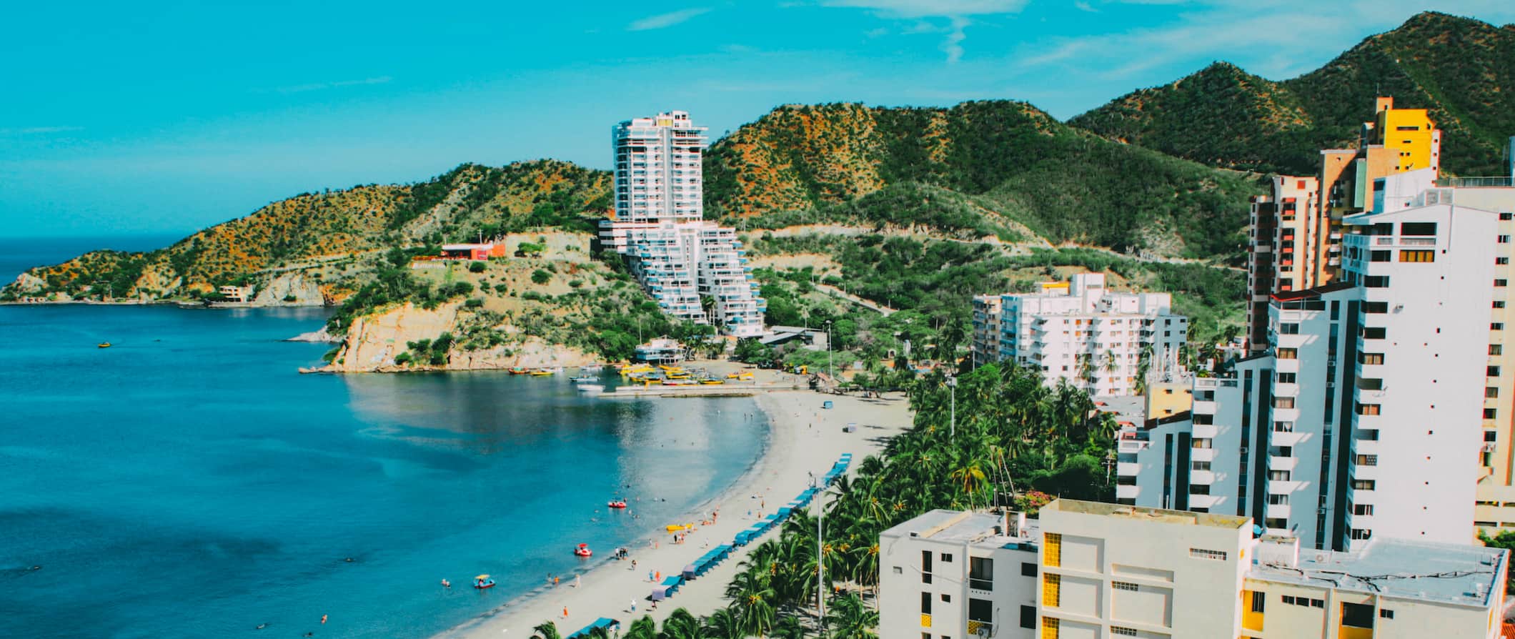 An aerial view of the beach front in Santa Marta, Colombia, featuring hotels along the coast and rolling hills in the background