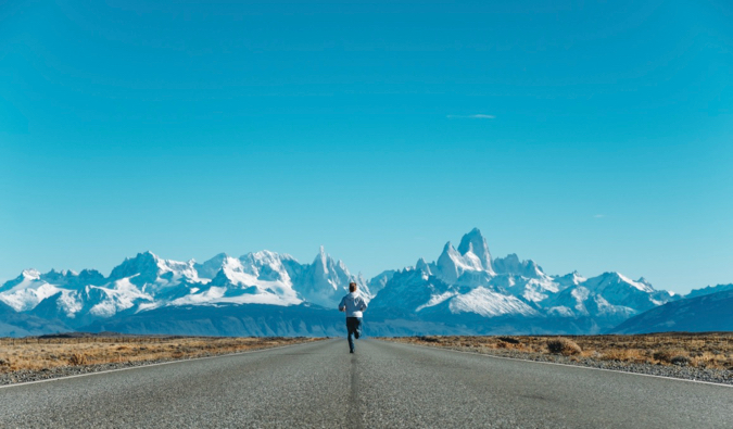 A solo traveler running down a paved road toward snow-capped mountains