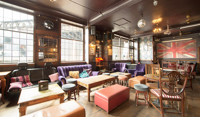 Cozy, spacious common room with lots of wooden tables and chairs, purple couches, and a large British flag on the wall at The Walrus Hostel, London