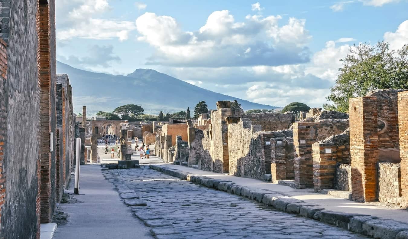 Cobblestone street in Pompeii, lined with ruins of buildings and Mount Vesuvius in the background.
