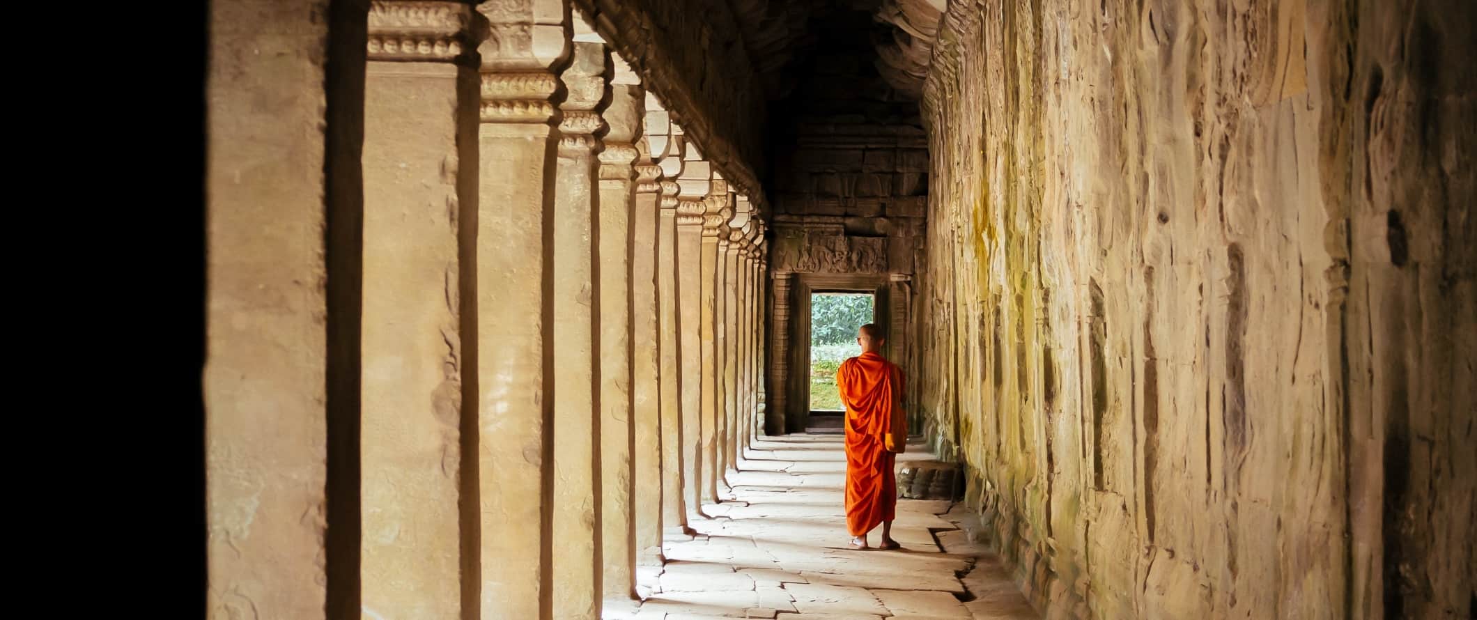 Monk in an orange robe walking down a temple's stone-lined walkway with stone pillars on one side at the Angkor Wat complex in Cambodia