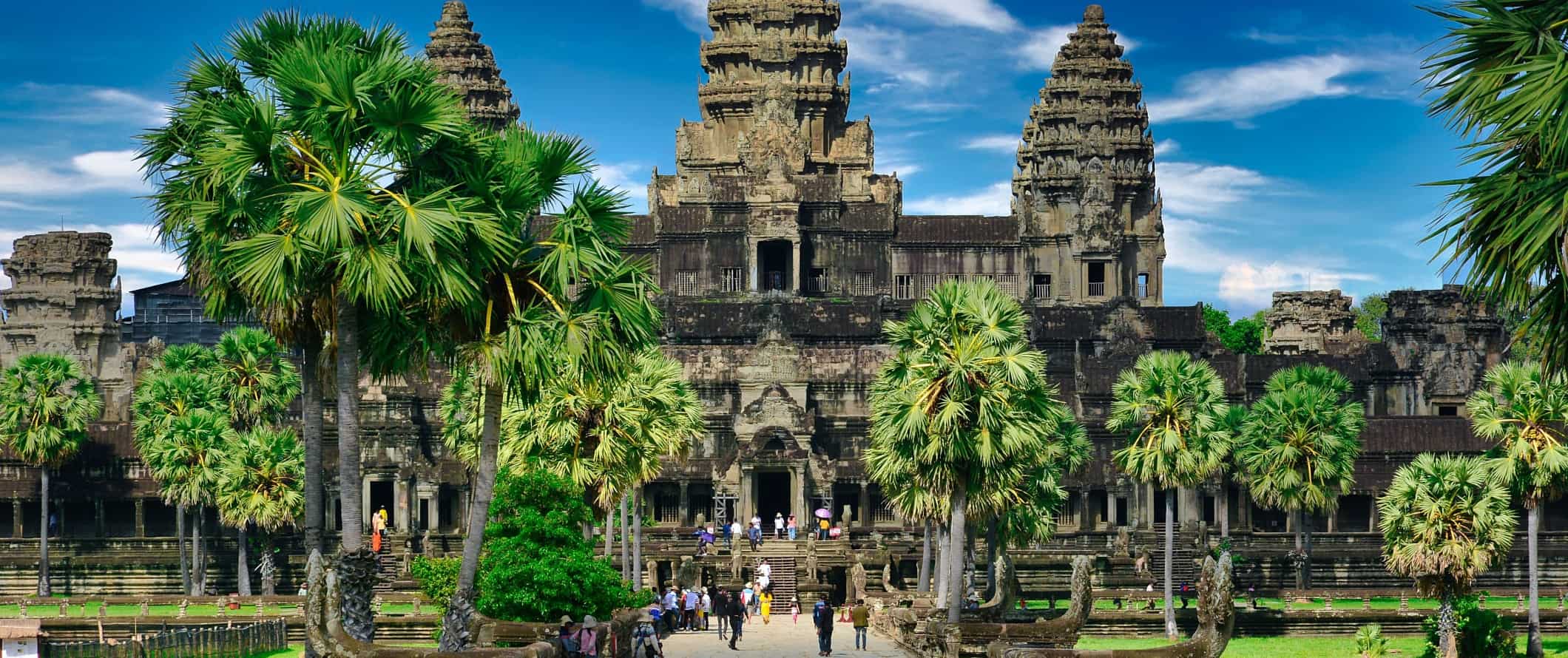 Visitors walking around in front of a large temple surrounded by tropical trees at the historic Angkor Wat complex in Cambodia