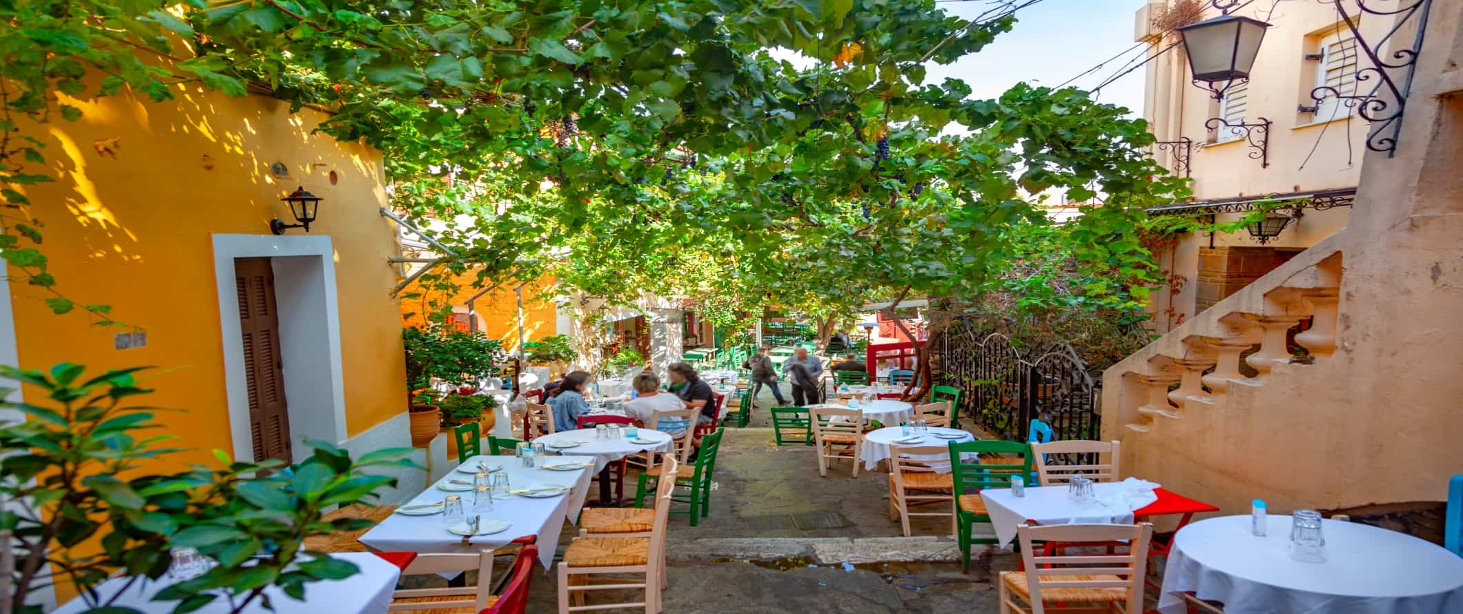 Cafe tables lining an alleyway covered with a lush tree branches, surrounded by bright yellow buildings, in Athens, Greece.