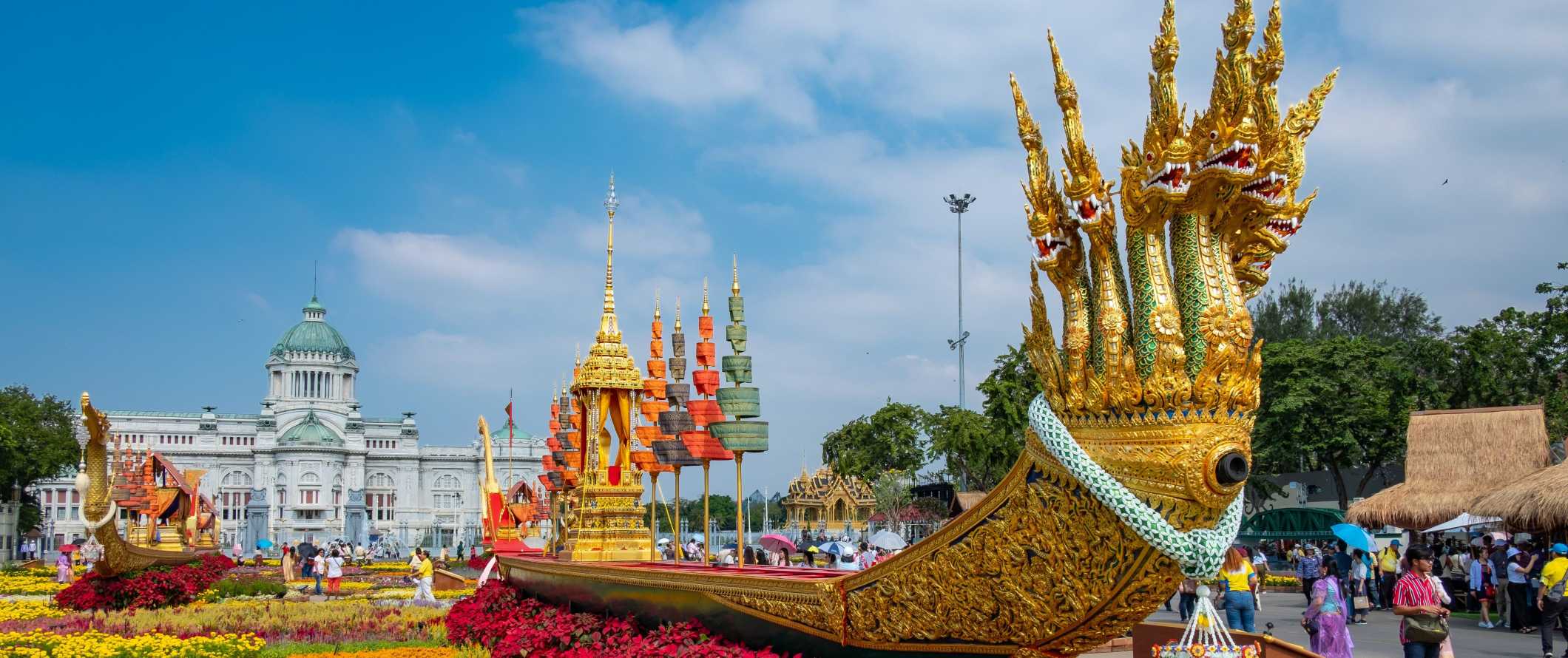 A large, long golden barge with many dragon heads sits on a bright flower bed in front of the Royal Barge Museum in Bangkok, Thailand