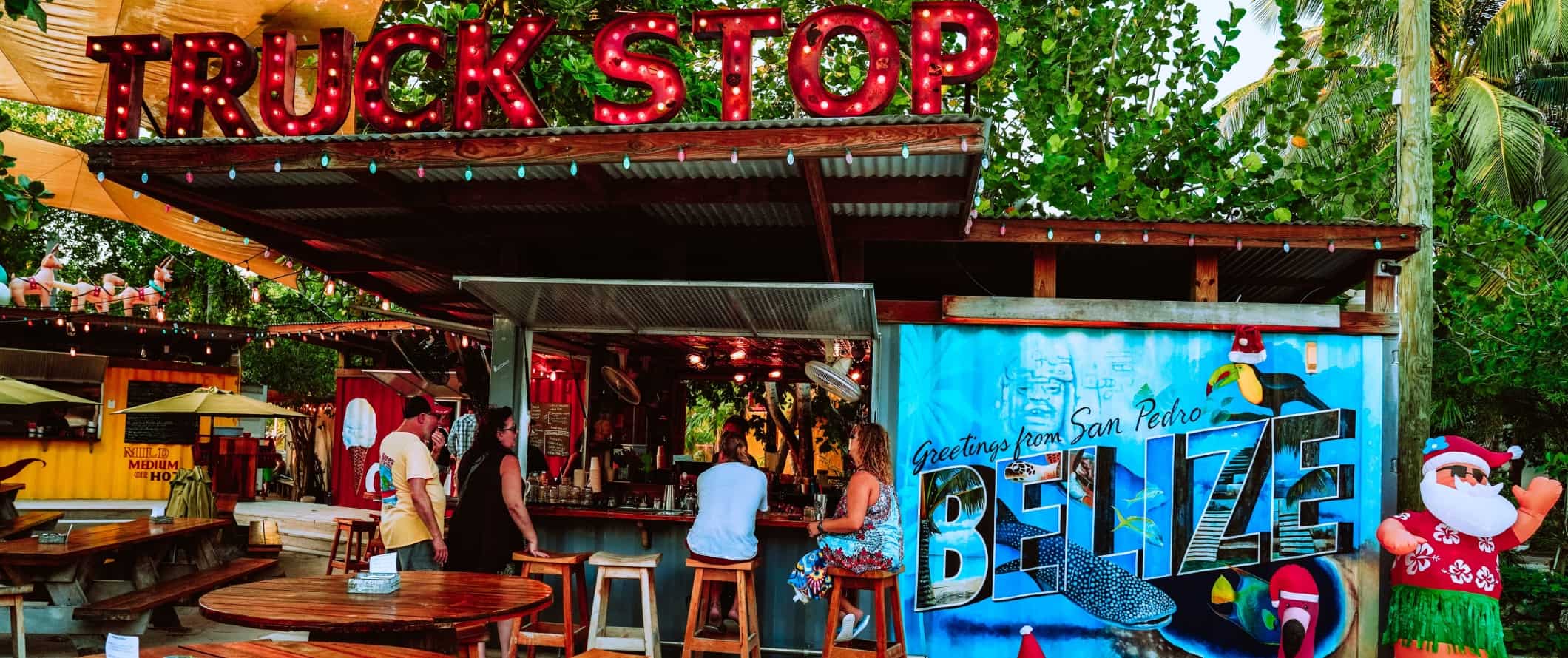 People sitting at a beach bar with a huge neon sign that says 'Truck Stop' and a bright mural that says 'Greetings from San Pedro, Belize' 