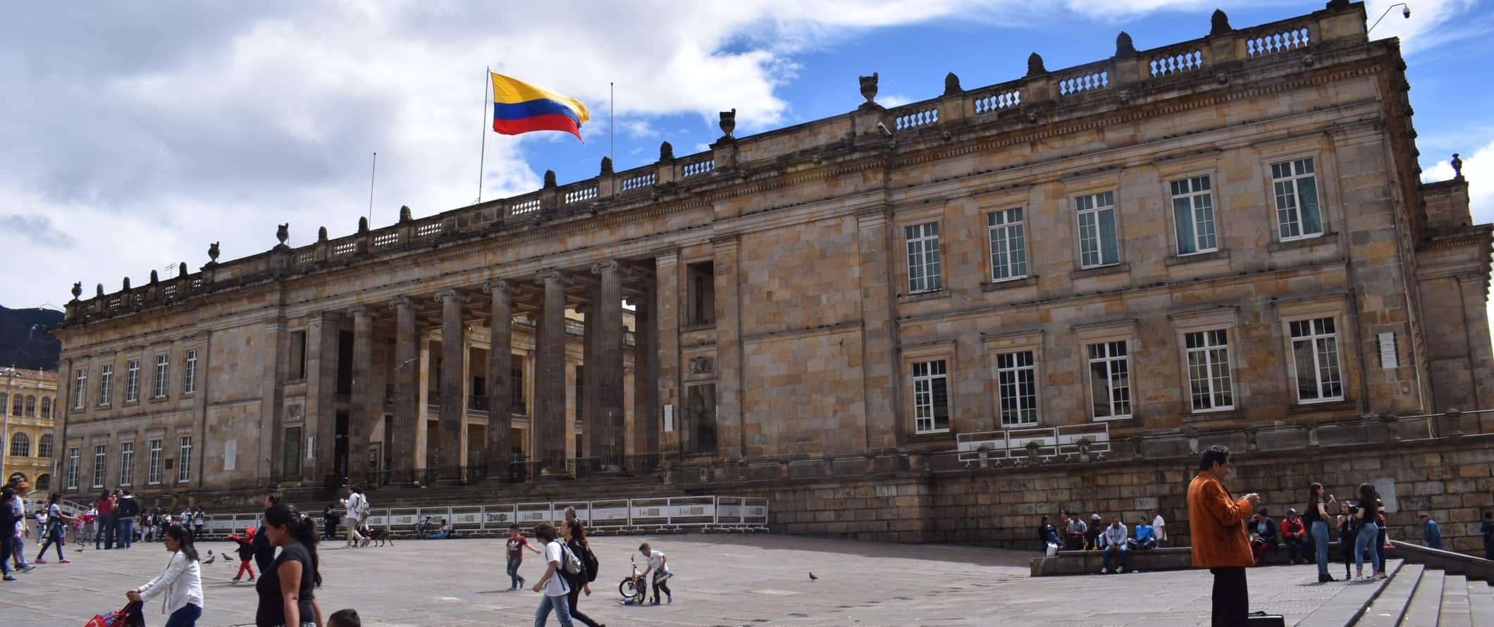 People walking around in front of a large neoclassical building with many pillars and the Colombian flag flying from the roof in Bogota, Colombia 