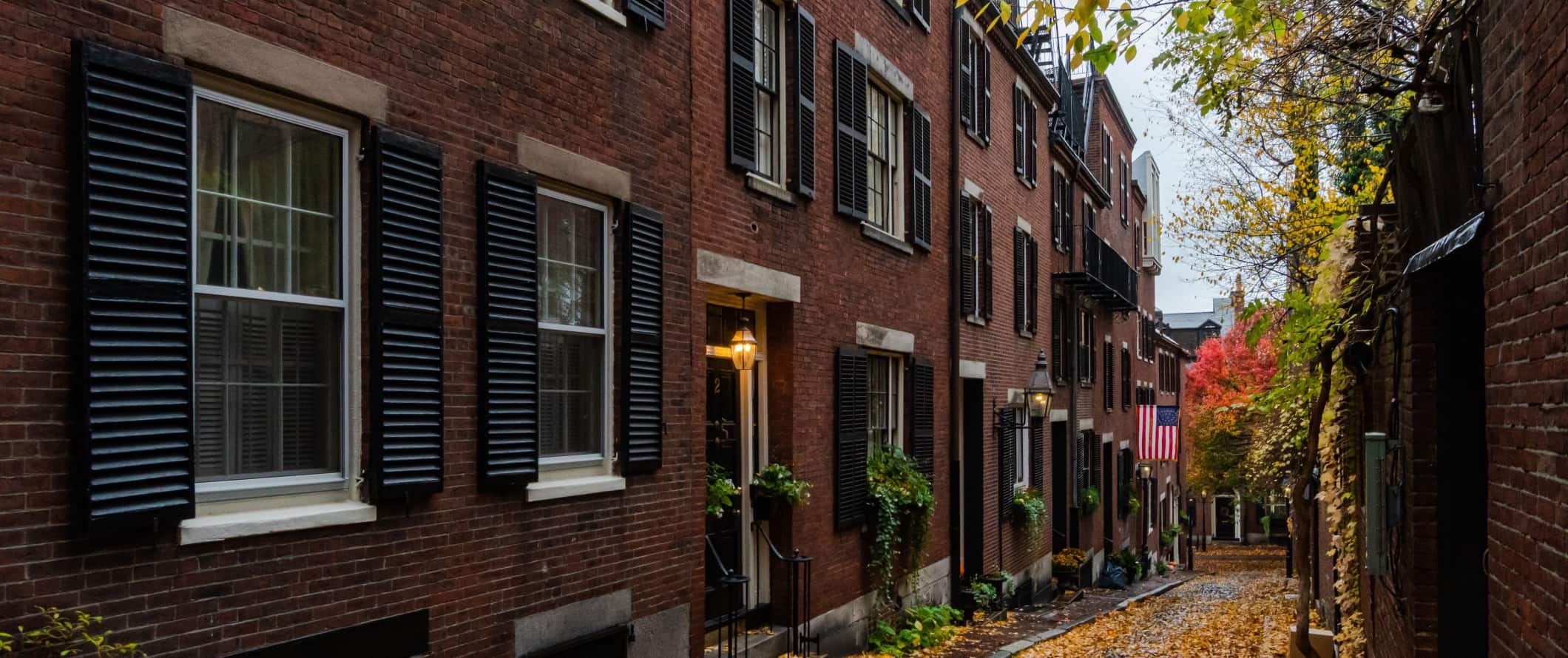 Historic brick houses with black shutters lining an alleyway with orange leaves on the ground in Boston, Massachusetts.