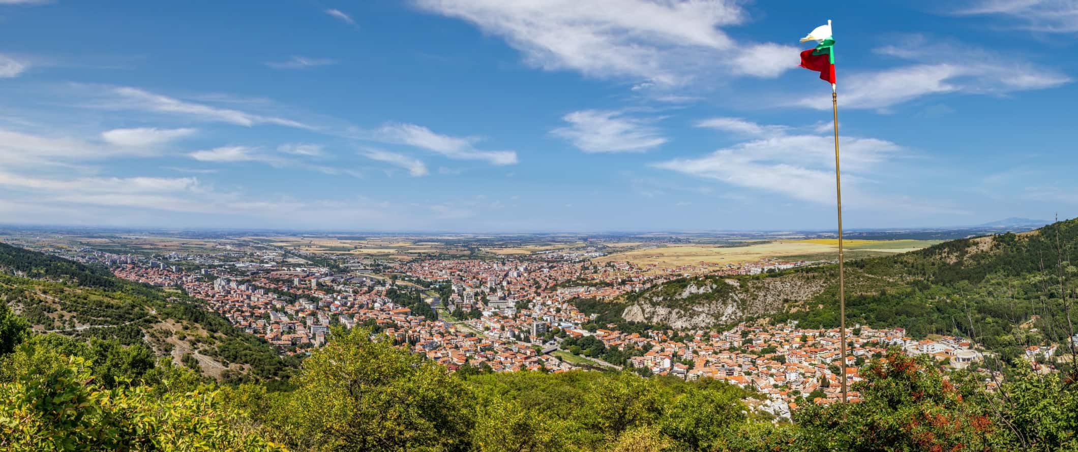 Panoramic views over the red terracotta rooftops of the historic town of Asenovgrad amidst rolling hills in Bulgaria, with the Bulgarian flag flying in the foreground