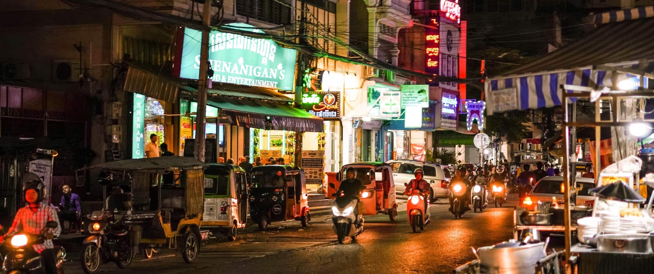 Mopeds, motorbikes, and tuk tuks driving along a street lit up at night in Phnom Penh, the capital city of Cambodia
