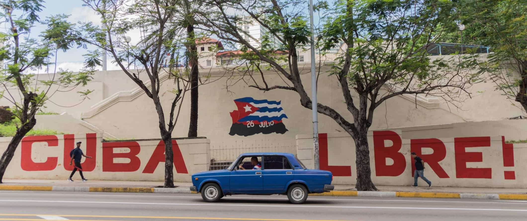 An old blue car driving down the street past a mural that says 'Cuba Libre' with a Cuban flag, in Havana, Cuba