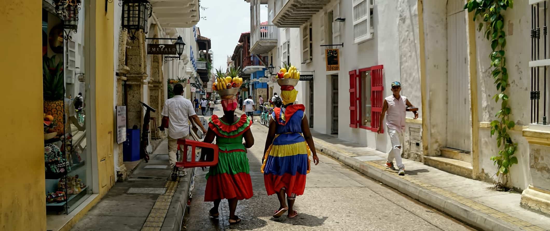 Two women in bright, colorful dresses, walking down a street with baskets of fruit on their heads in Cartagena, Colombia