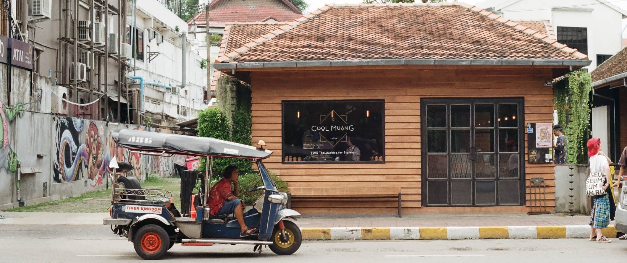 Person driving a rickshaw past a coffee shop as people walk down the street in Chiang Mai, Thailand
