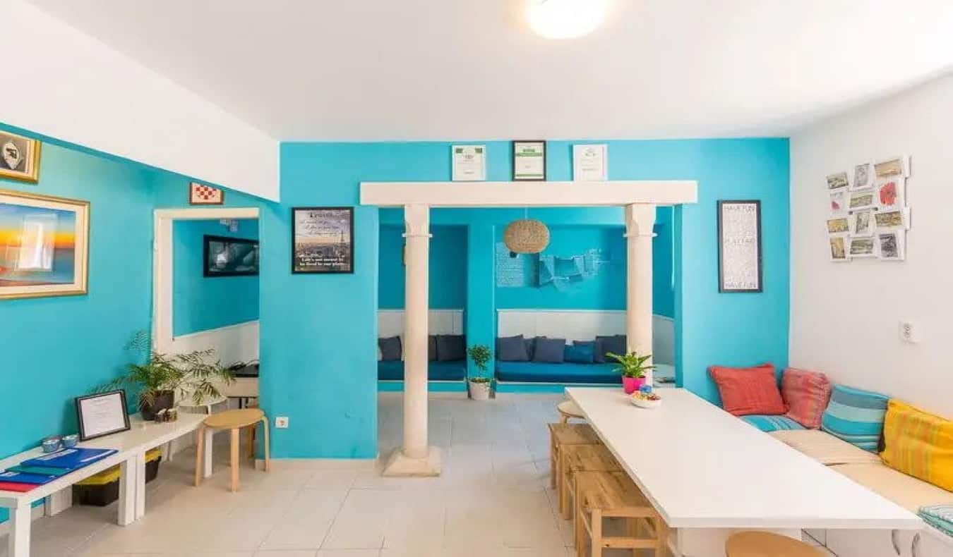 Bright common area with light blue walls, a long table, and bench with pillows at City Walls Dubrovnik hostel in Dubrovnik, Croatia.