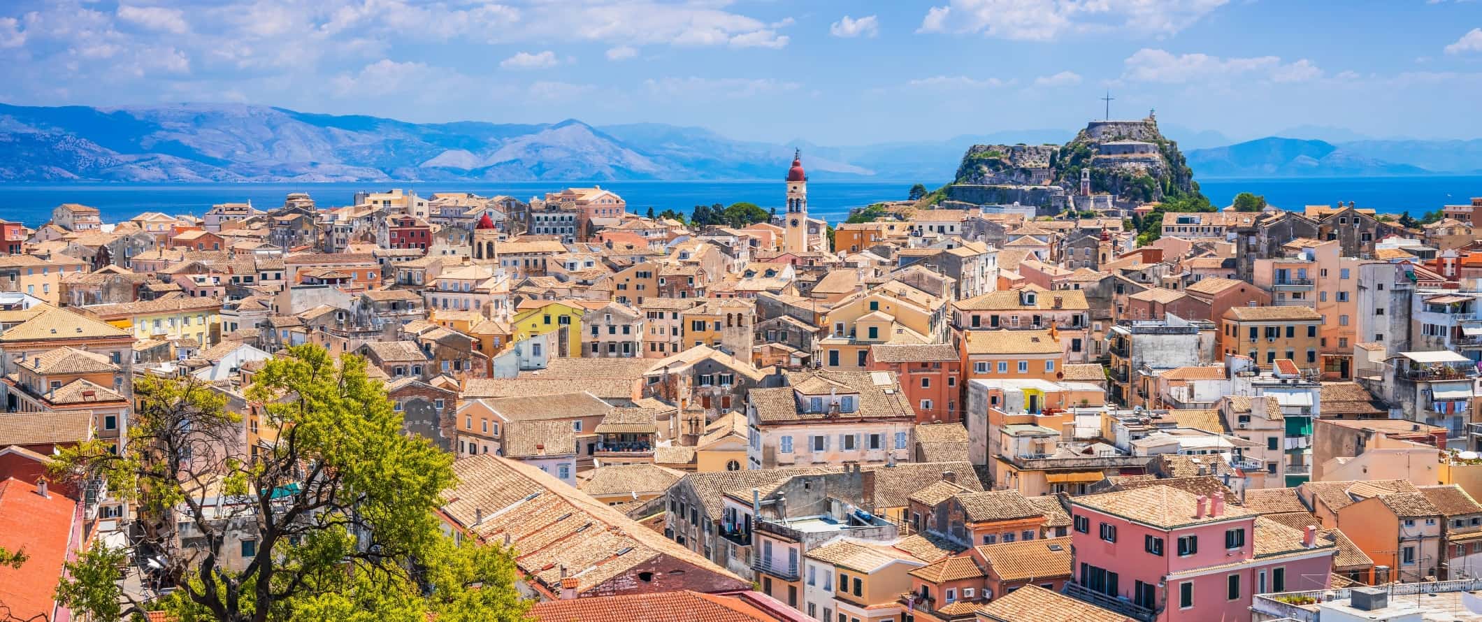 Aerial view of Corfu Town in Corfu, Greece, showing brightly colored houses with tiled roofs, and oceans and mountains in the background.