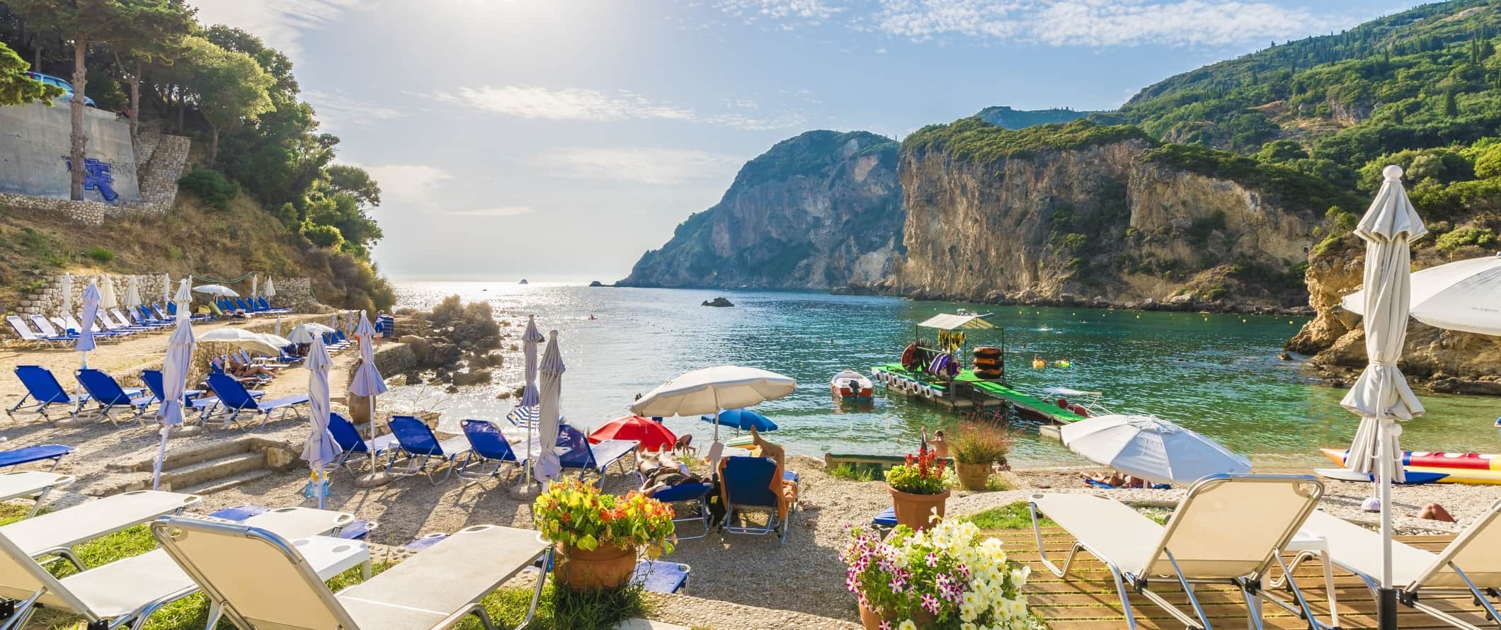 Beach loungers on a beach with rocky, tree-covered cliffs in the background and clear, turquoise waters in Corfu, Greece.