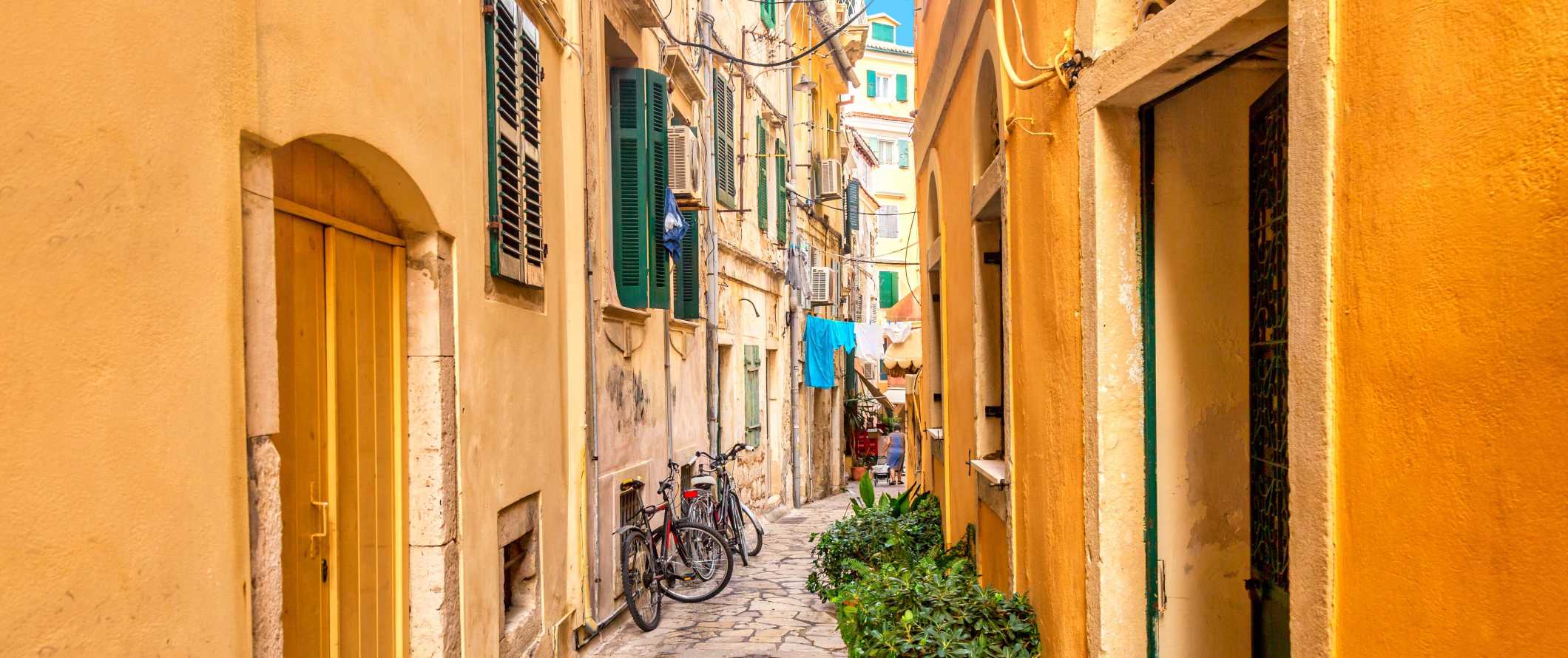 Small, flagstoned street lined with bright yellow buildings with green shutters in Corfu, Greece.