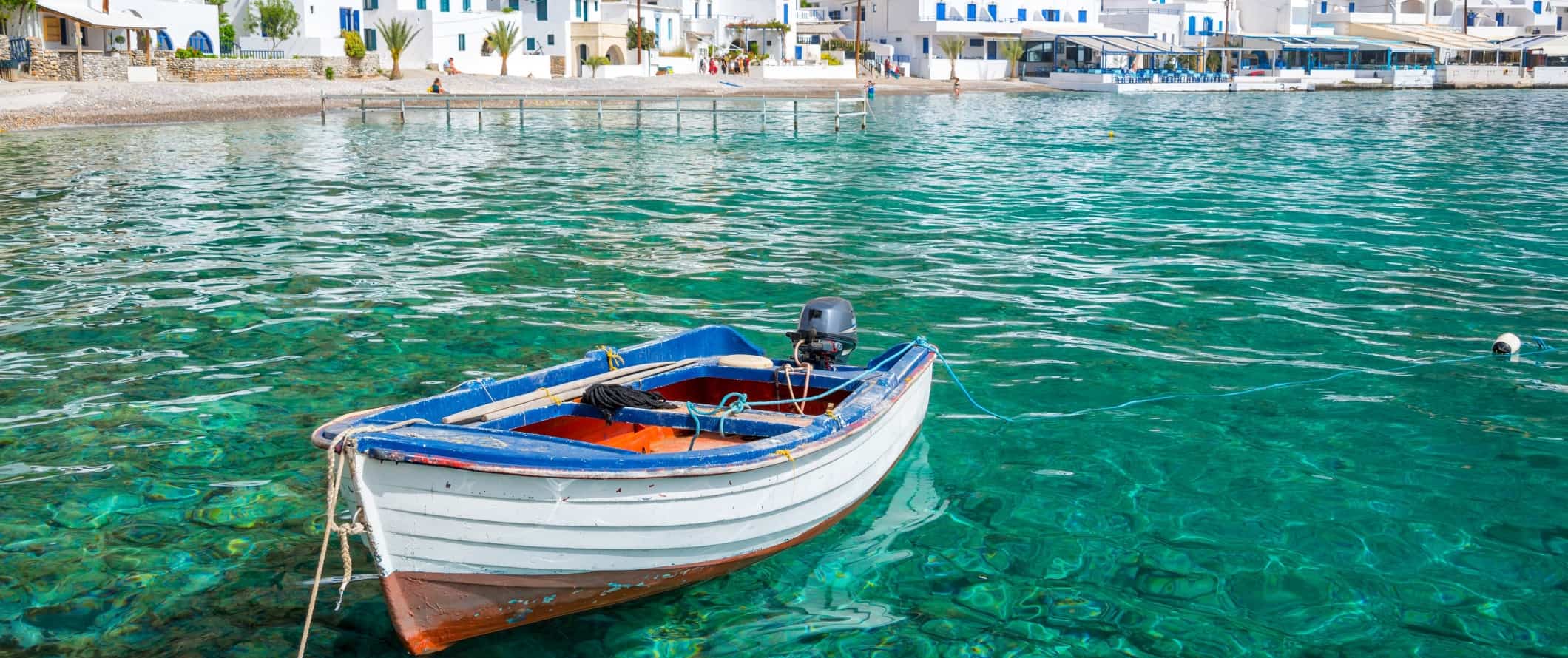 Lone boat in the clear turquoise waters of a bay lined with white houses on the island of Crete in Greece.