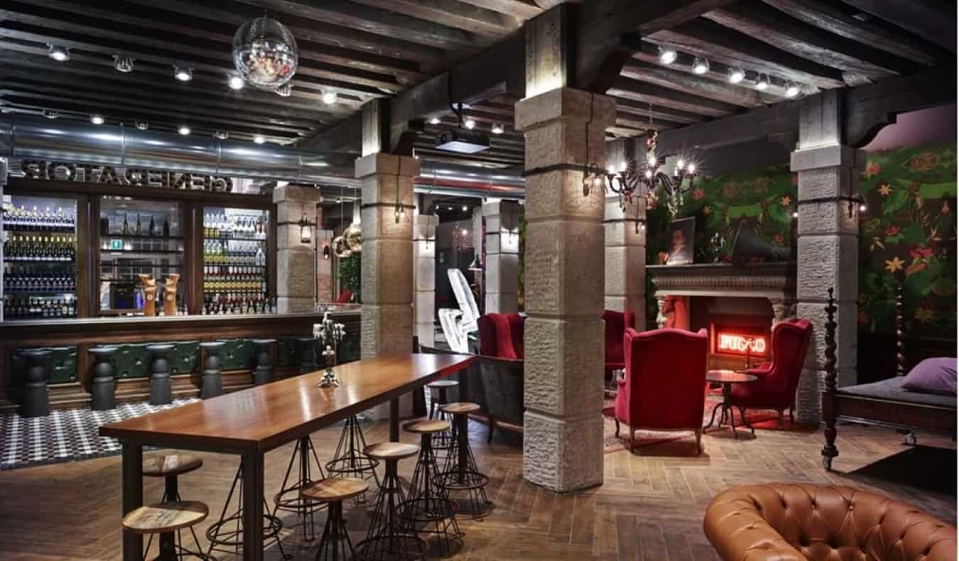 A common area with a bar, bar tables and cozy red chairs next to a fireplace at Generator Hostel in Venice, Italy.