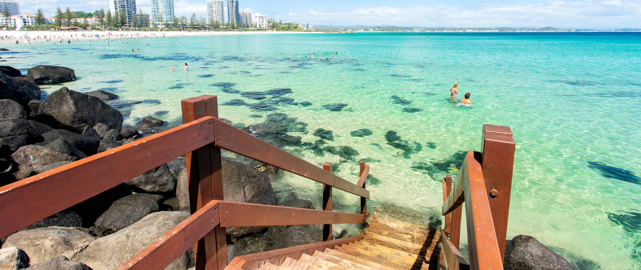 People swimming in the clear waters of Gold Coast, Australia near a small wooden staircase with the city in the background