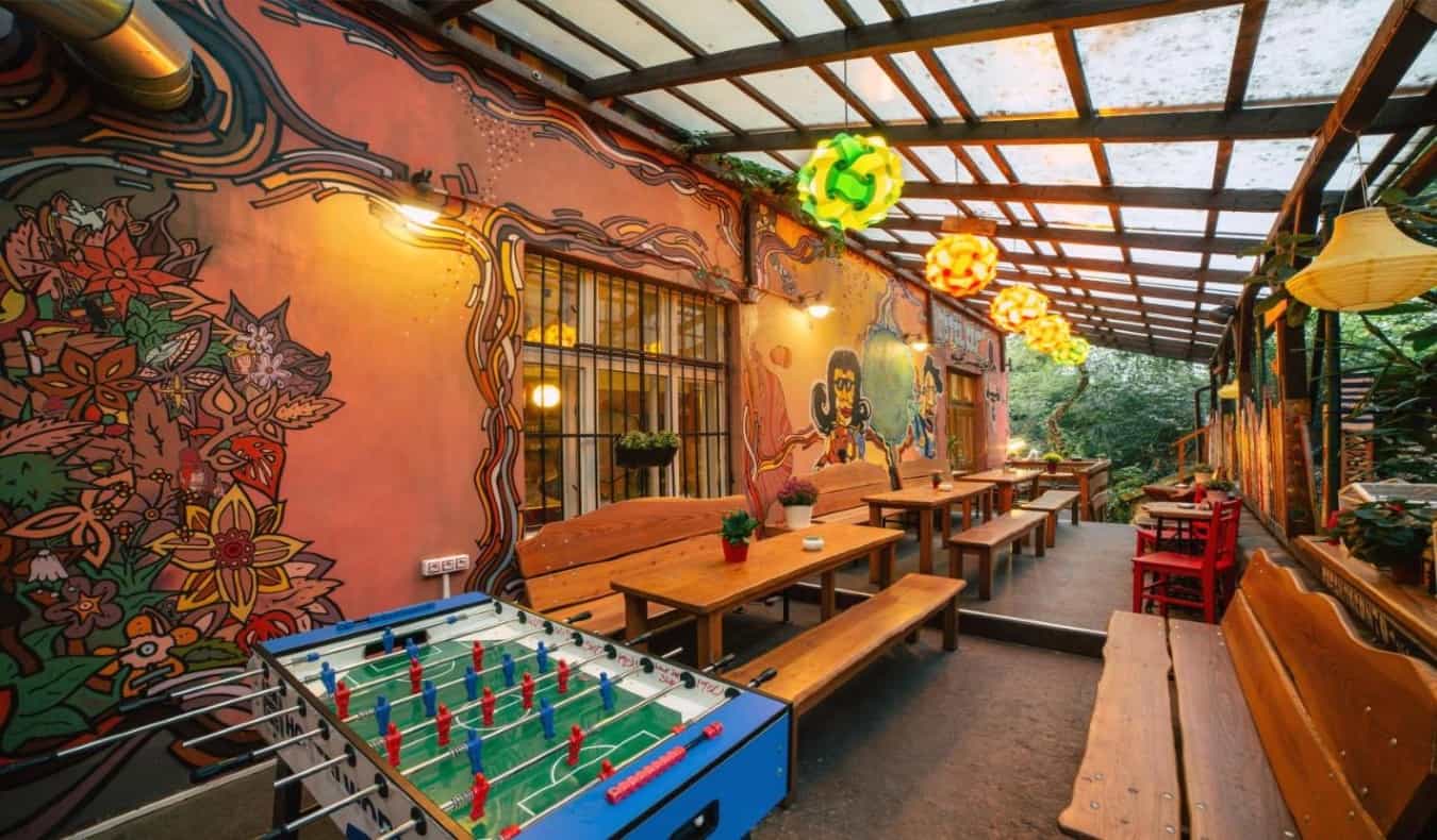 covered outdoor beer garden with long wooden picnic tables and benches, a foosball table, and murals painted on the building at Hostel Elf in Prague.