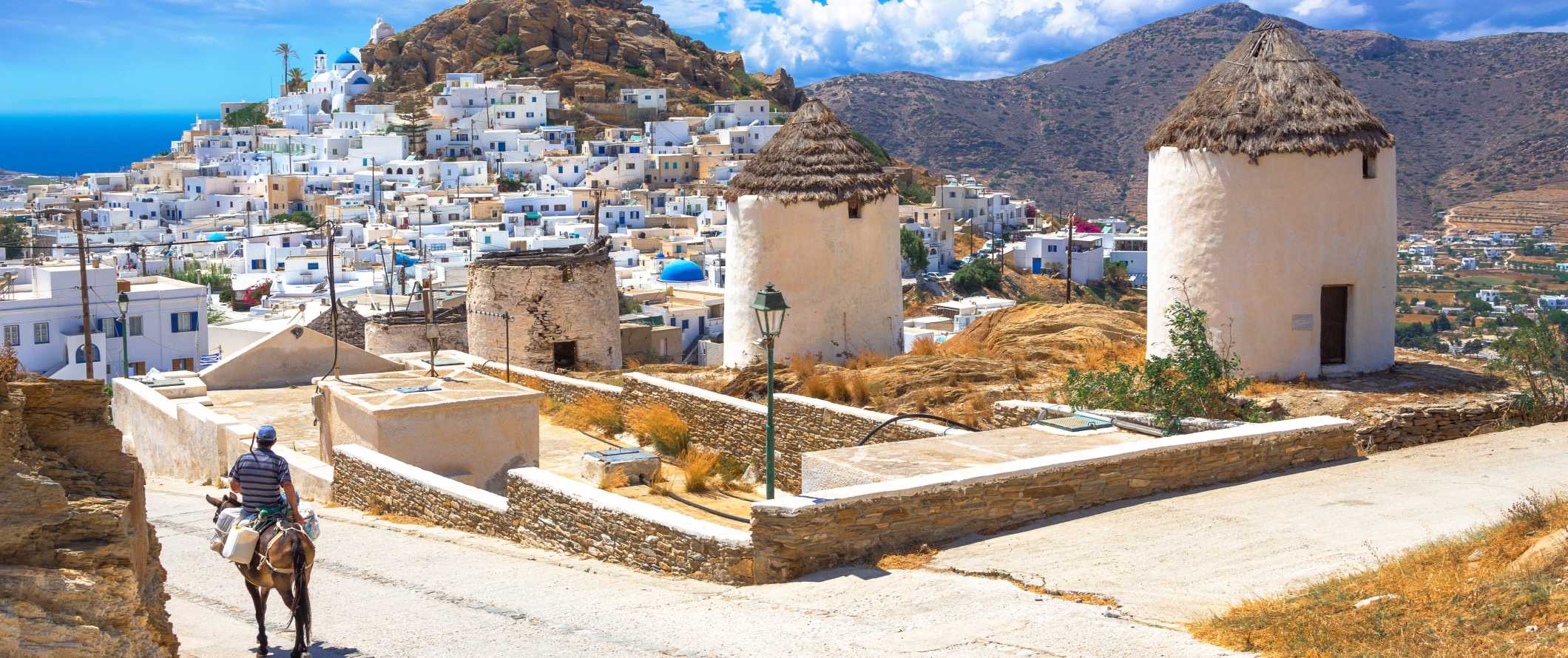 View of Chora with white-washed houses, windmills in the foreground, and a man riding a donkey down the road in Ios, Greece