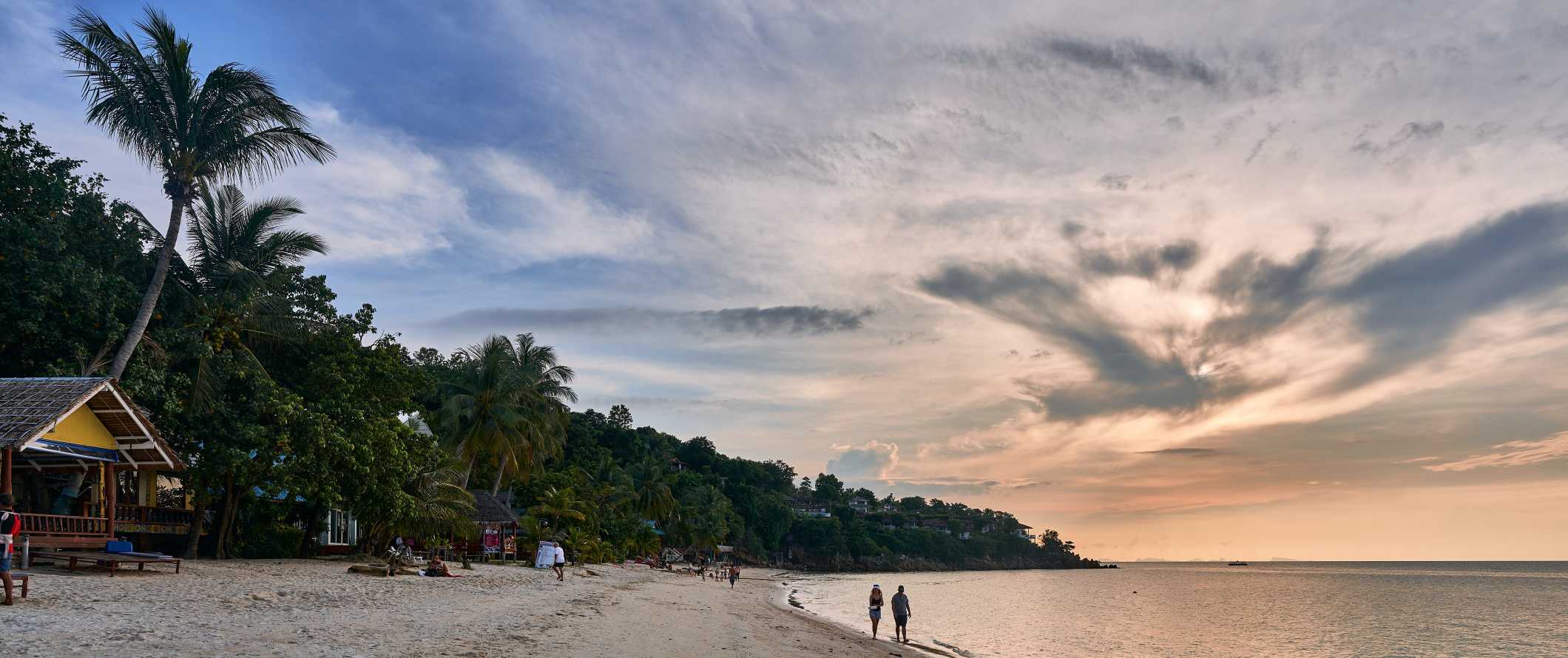 People walking along a beach at sunset on the island of Ko Pha Ngan, Thailand at sunset over the ocean
