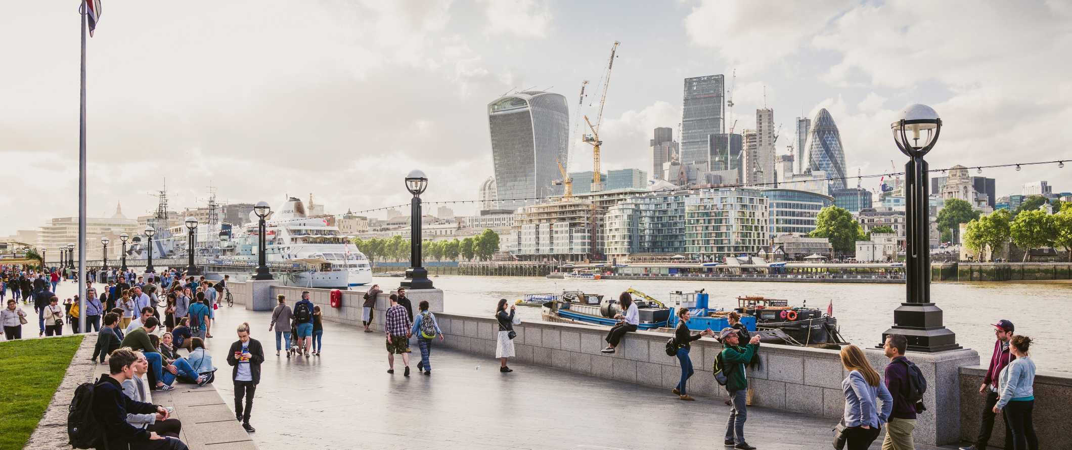 People walking around on the waterfront of the River Thames with London's iconic skyscrapers in the background