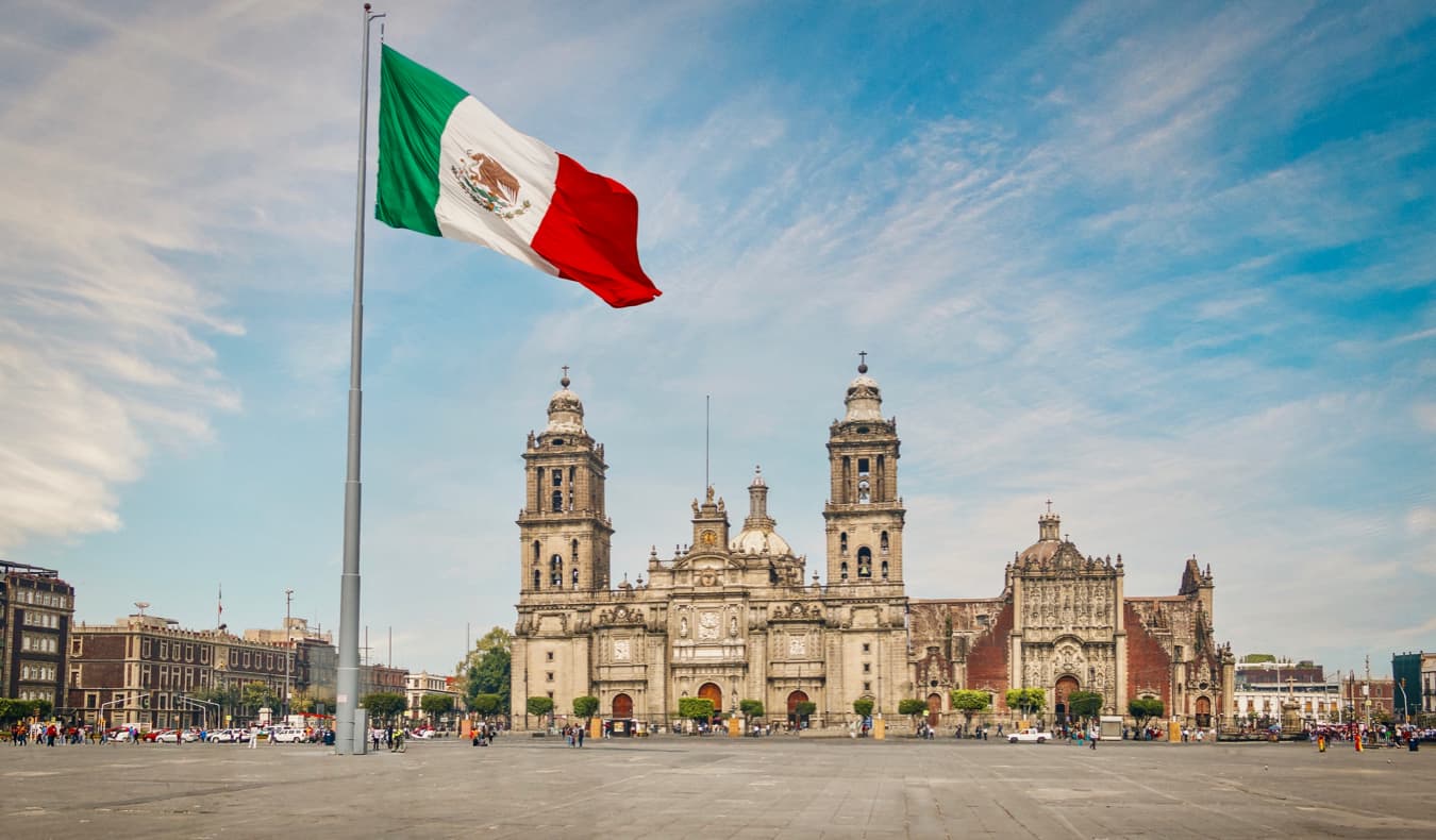 A large Mexican flag in front of several historic buildings in Mexico City