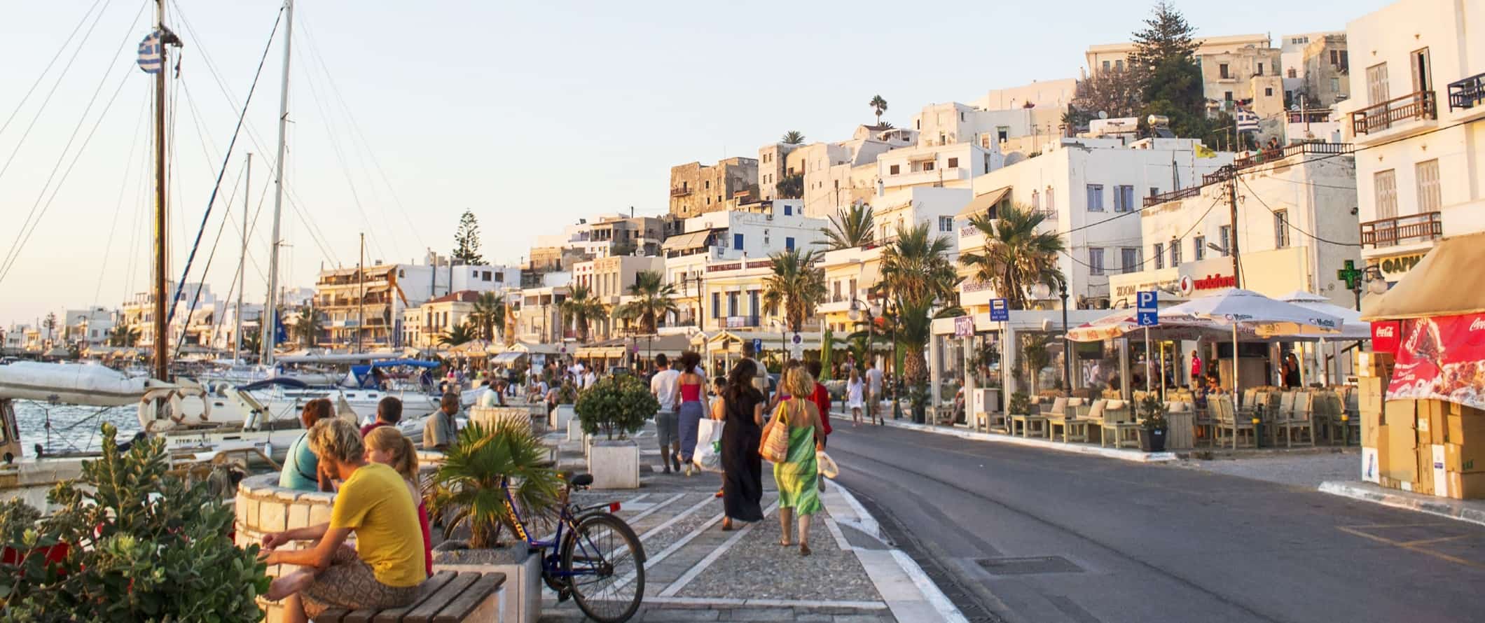 People sitting on benches and walking along the harborfront filled with sailboats, and Chora Old Town with its whitewashed buildings in the background on the island of Naxos in Greece.