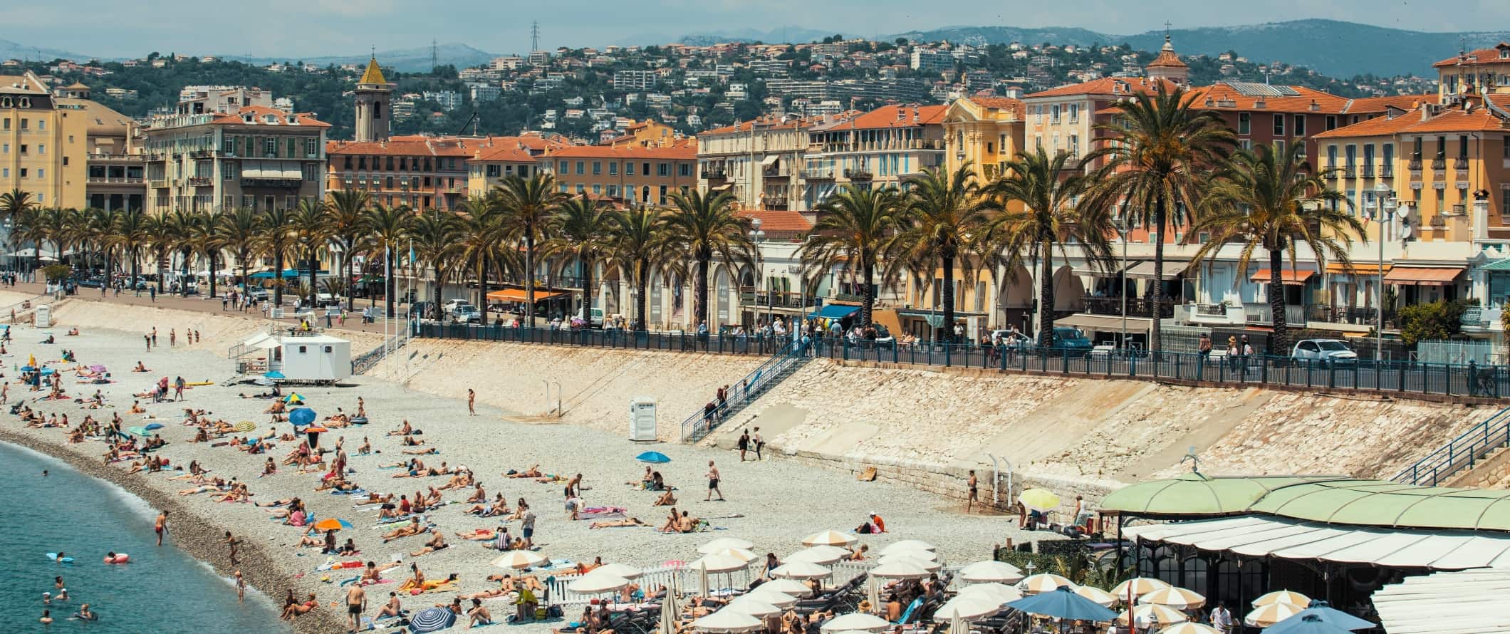 People lying on the beach in front of a palm-tree-lined promenade with the city of Nice, France rising in the background