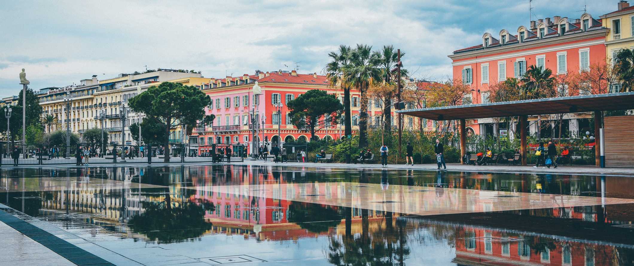 A wide fountain and reflecting pool with the colorful buildings of Nice, France in the background on a dreary day
