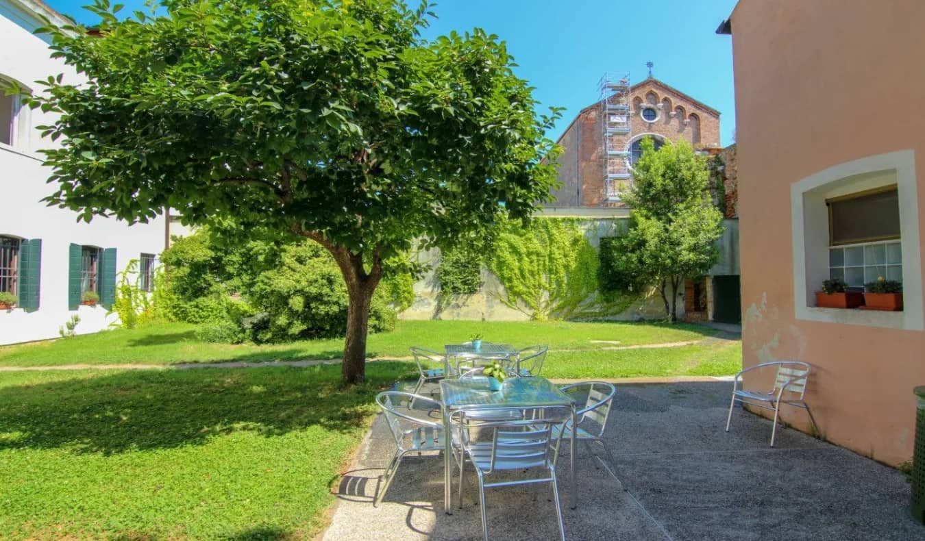 A courtyard surrounded by trees, metal tables and a church in the distance from the Ostello S. Fosca guesthouse in Venice, Italy.