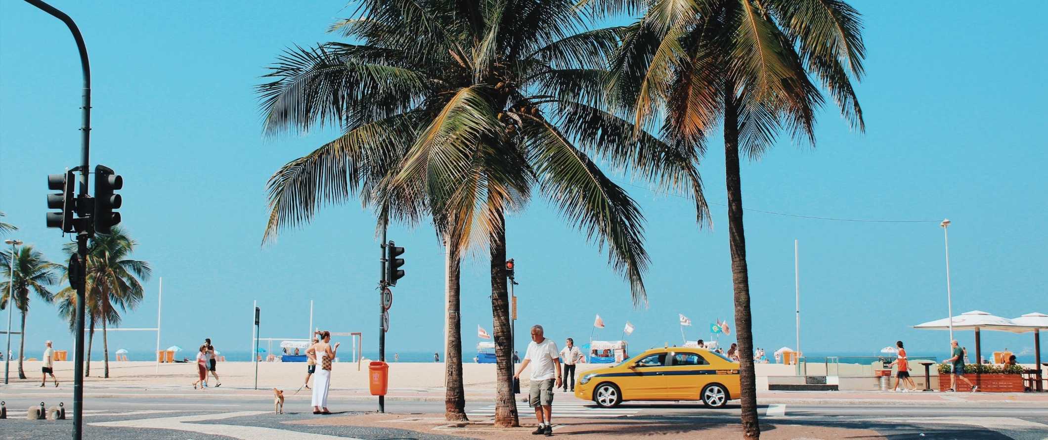People walking down a palm tree-lined street with a yellow taxi going by along the beach in Rio de Janeiro, Brazil