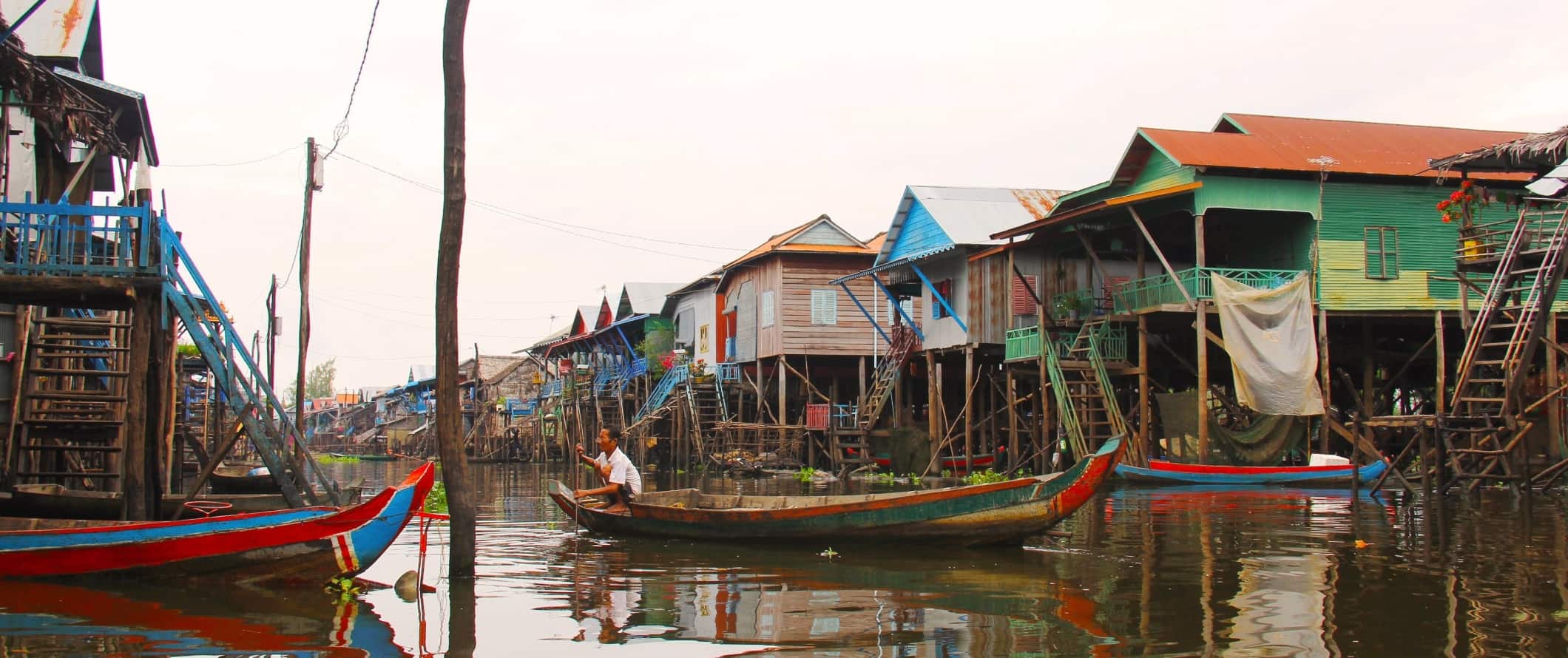 A man using a pole to maneuver a long boat down a waterway surrounded by colorful houses on stilts in Tonle Sap, Cambodia
