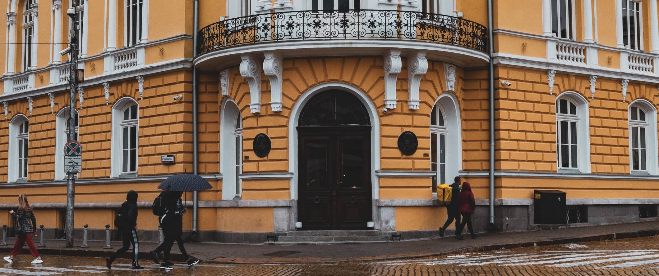 People walking down the street in the rain, past a bright yellow building in Sofia, Bulgaria