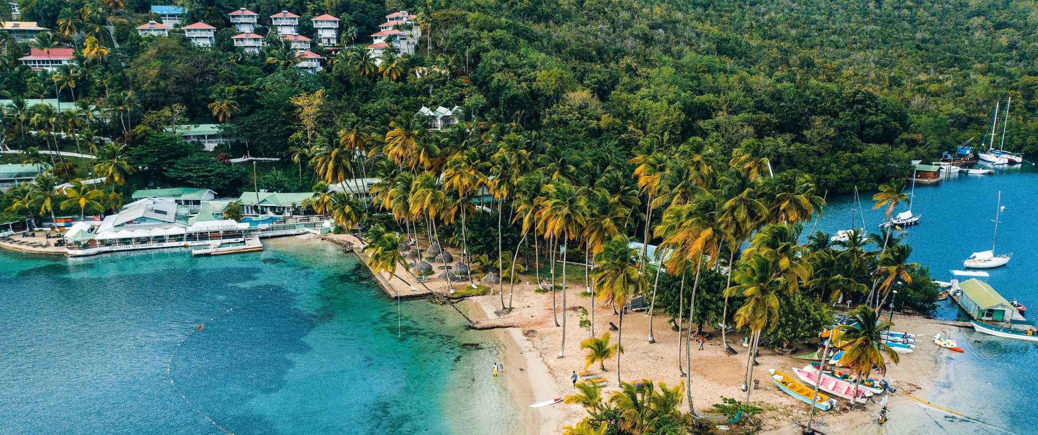 Drone view of a small turquoise bay with boats along the palm-tree-lined beach and houses tucked into the lush hillsides of Saint Lucia