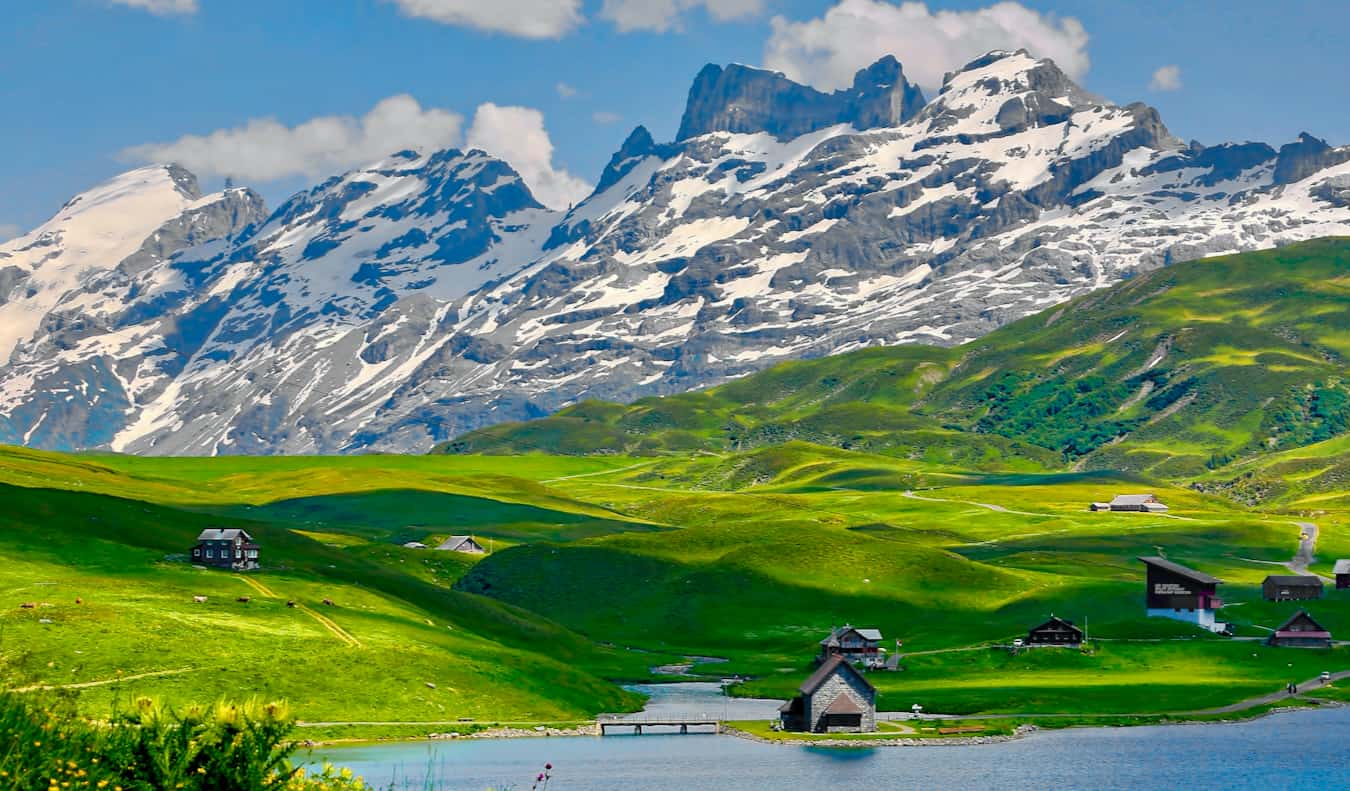 A scenic rural scene in Switzerland featuring small cabins on a lush, green hill near a snowy mountain