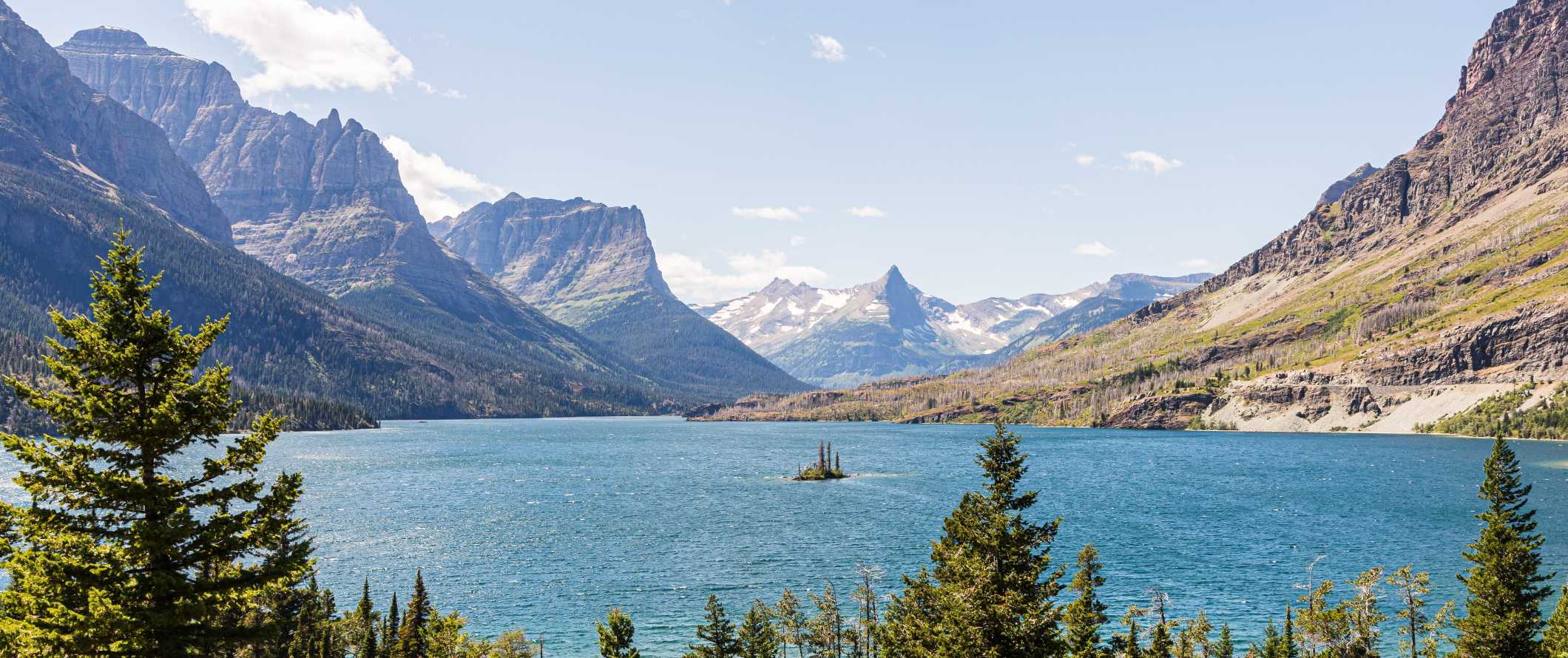 Dramatic, jagged mountains in front of an expansive blue lake with a small, tree-covered island in the middle, in Glacier National Park, United States.