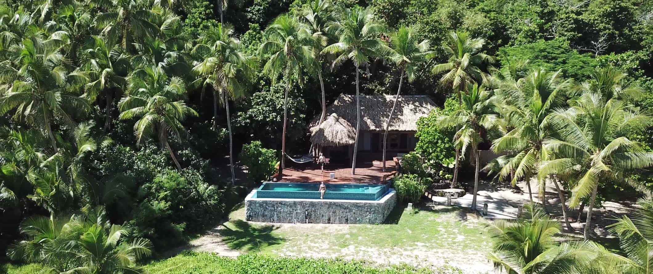 People sitting at a private pool in front of a thatched-roof hut surrounded by palm trees in the Yasawa Islands in Fiji