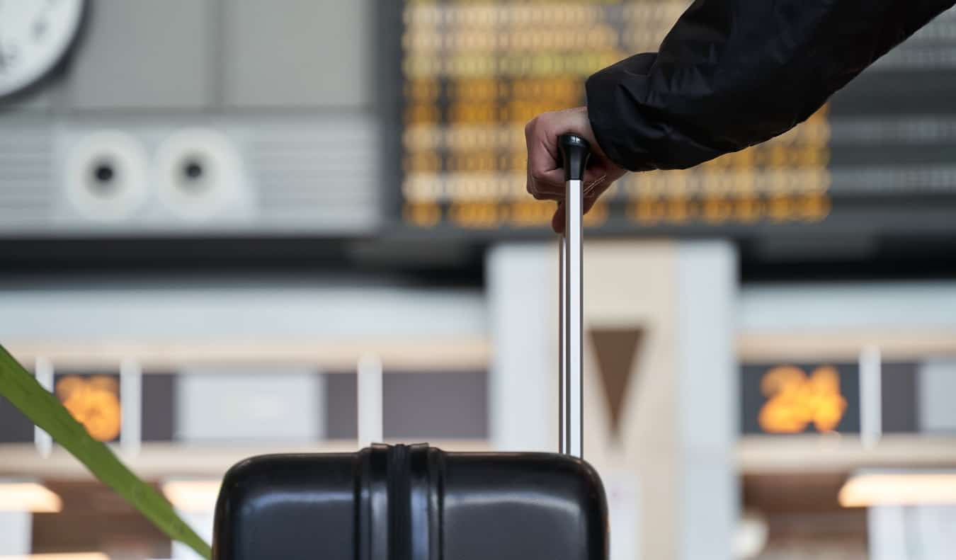 Person holding onto the handle of a rolling suitcase while looking at an airport departures board