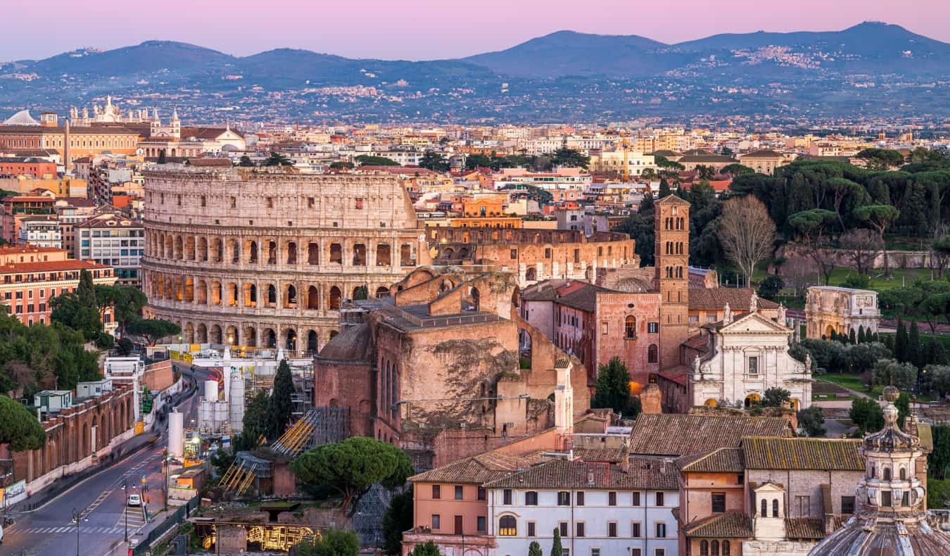 The skyline of Rome, Italy at sunset, with historic buildings including the Colosseum in the foreground and mountains in the background