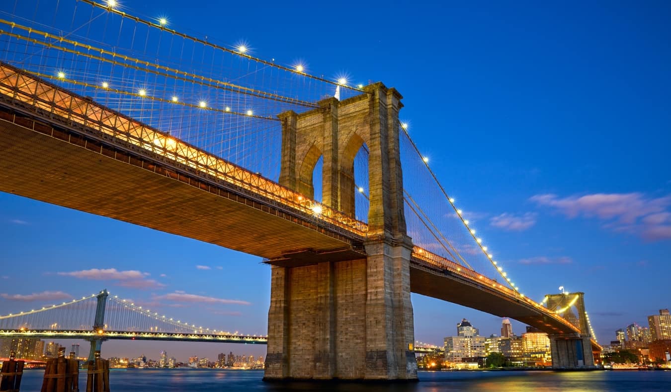 Full span of the Brooklyn Bridge in front of the Manhattan skyline lit up at night in New York City, USA.