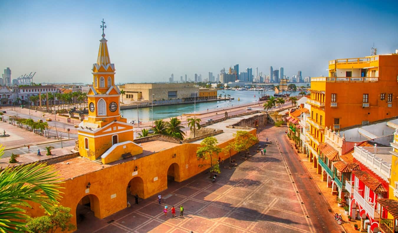 A view over an expansive plaza surrounded by bright orange historic buildings with the harbor and modern skyscrapers in the background in Cartagena, Colombia