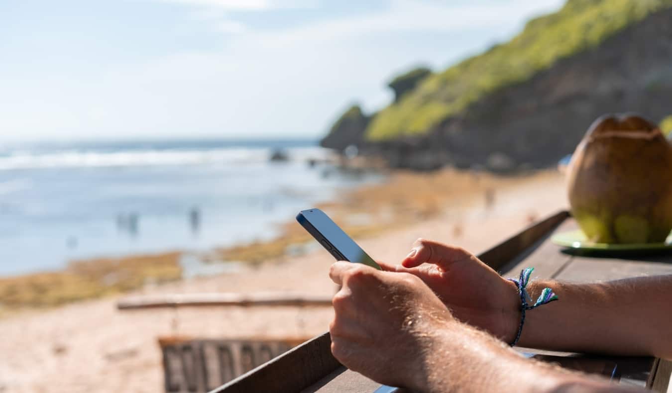 Close up on a man's hands holding a cell phone with a coconut and beach in the background