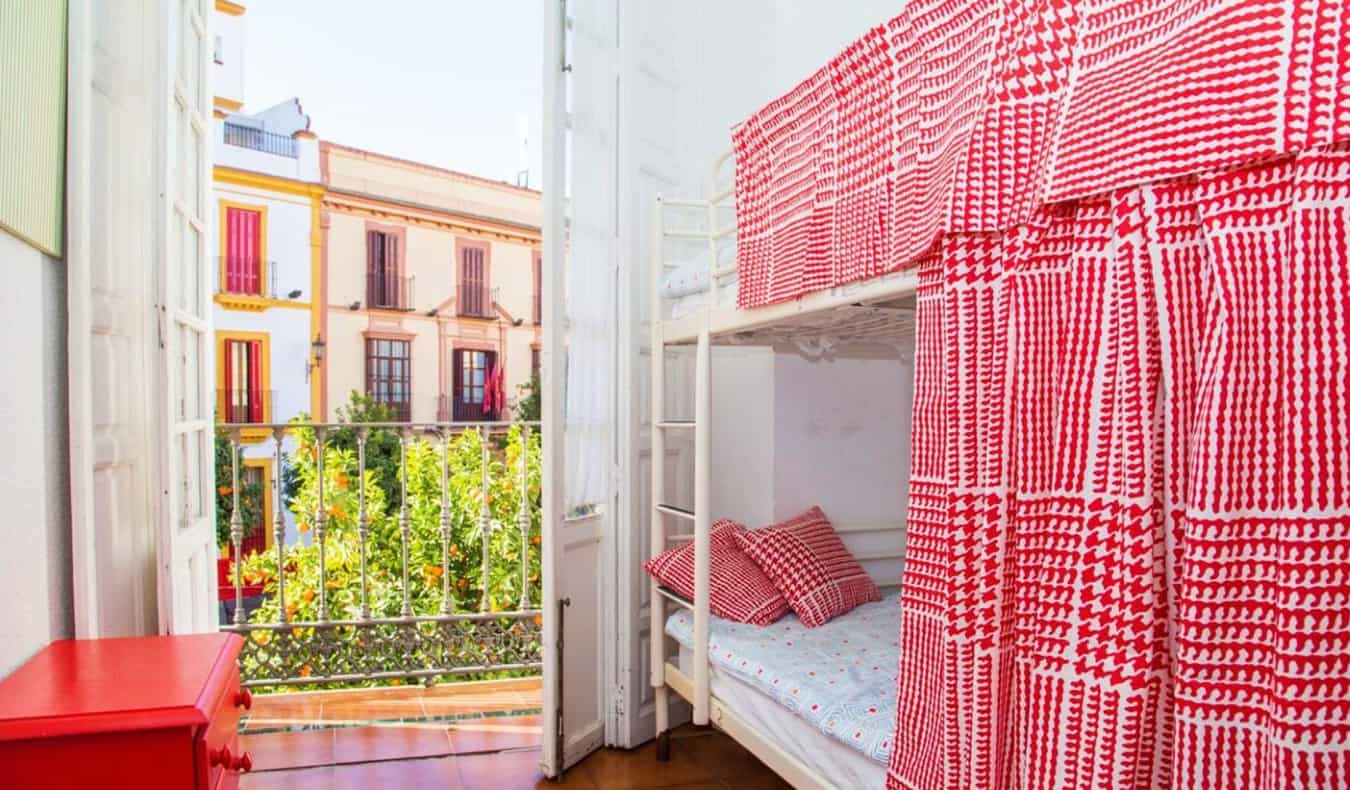 Bunk beds with red polychrome privacy curtains, with an unshut door to a terrace showing the brightly colored buildings of Sevilla, Spain in the background