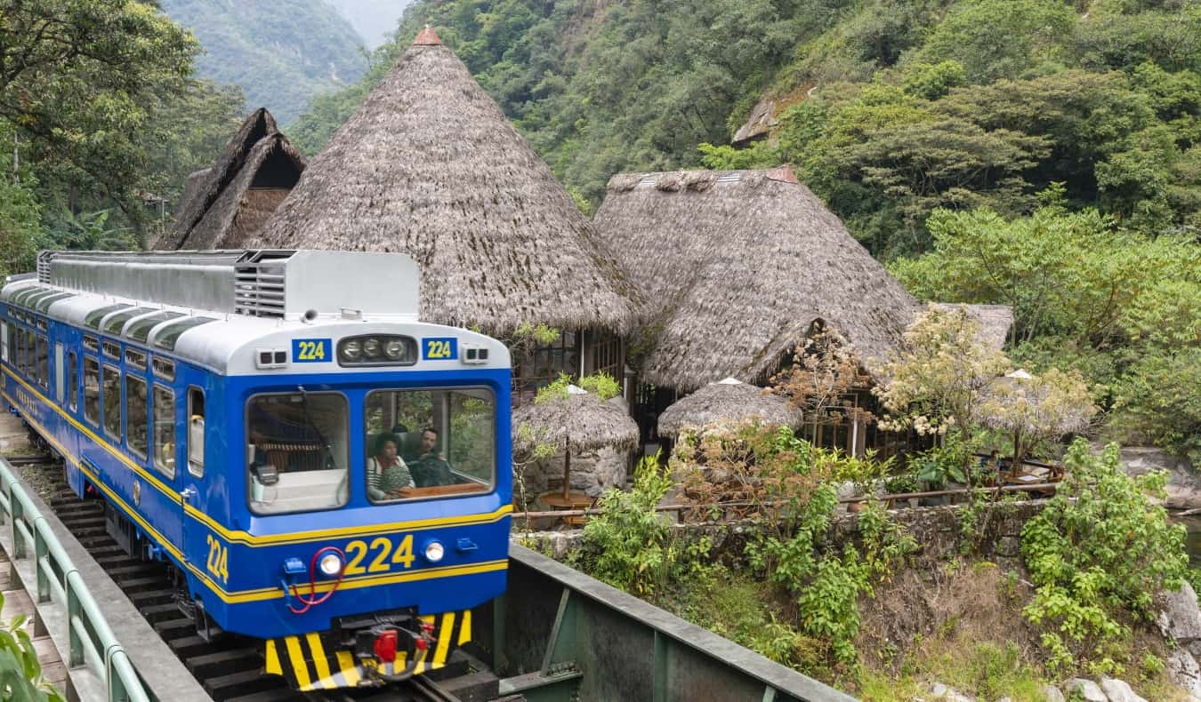 An old blue train passes by thatched houses near Machu Picchu in Peru