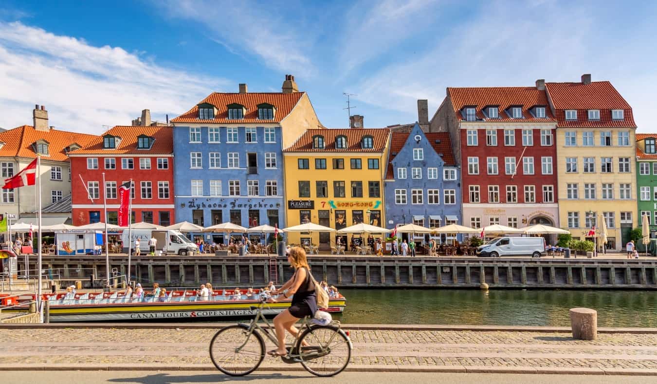 A woman biking by colorful, historic buildings lining a canal in Indry By, the historic center of Copenhagen, Denmark