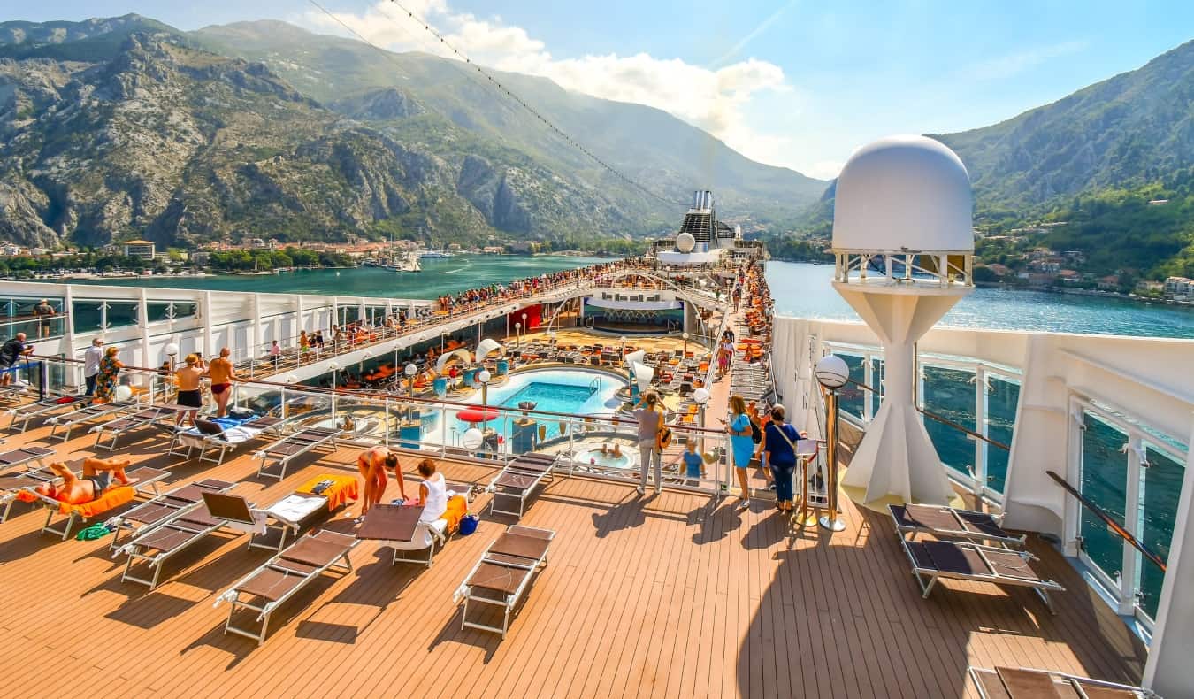 Cruise deck with people lounging on chairs next to a pool as the ship pulls into the bay of Kotor with green rolling mountains in the background