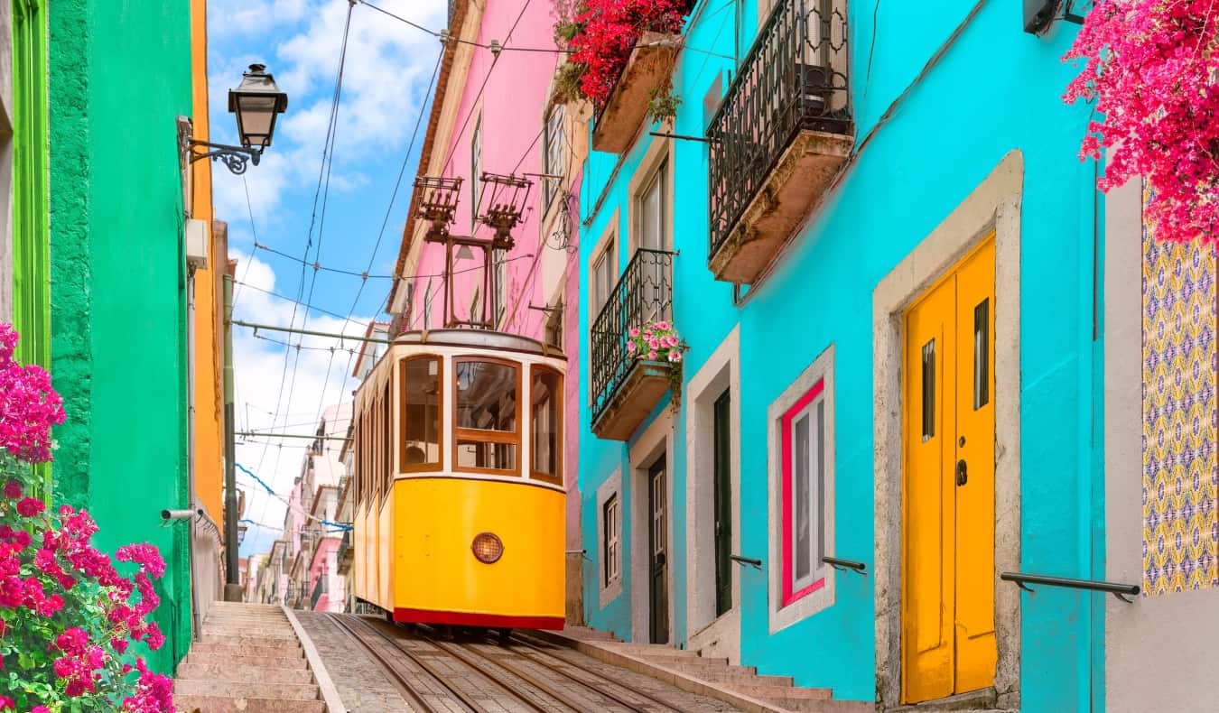 Historic yellow tram on a street going down a hill lined with colorful buildings and flowers on the balconies in Lisbon, Portugal